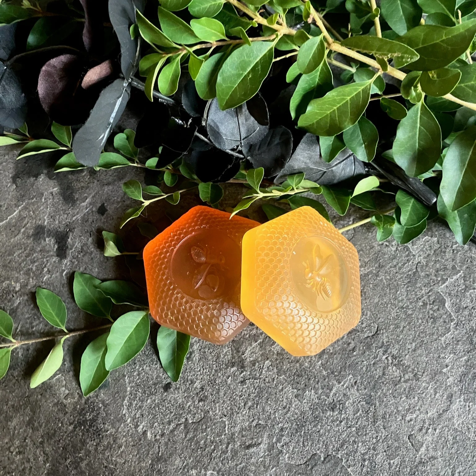 Two hexagonal honeycomb soaps, one dark amber and one pale gold, on slate with green leaves